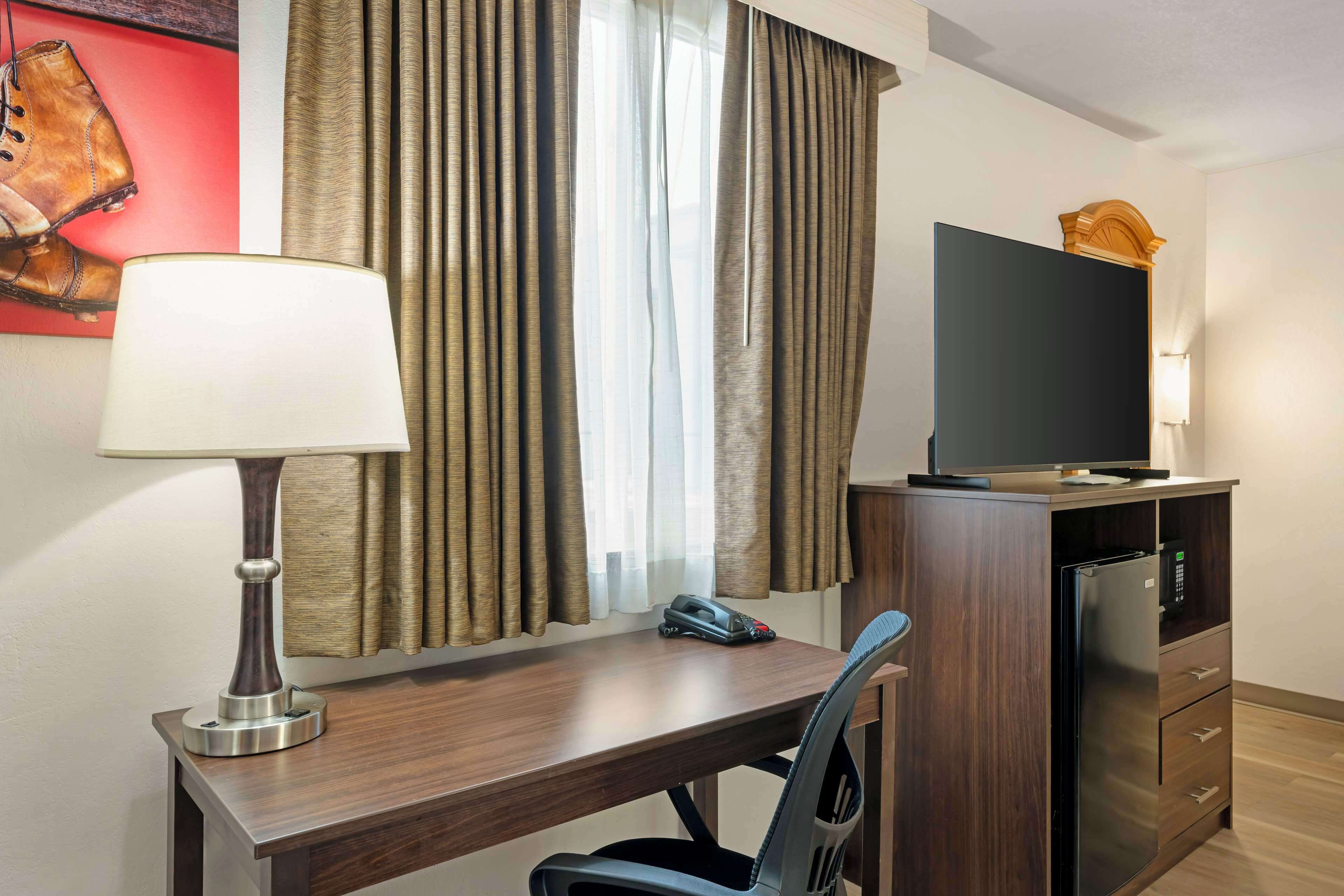 A hotel room with a desk, a chair, a television, a refrigerator, and a lamp.
