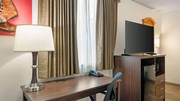 A hotel room with a desk, a chair, a television, a refrigerator, and a lamp.