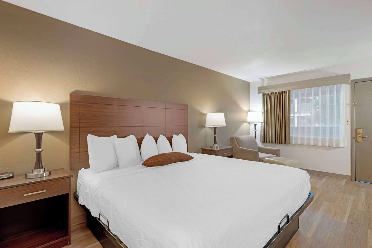 Une chambre d’hôtel avec un grand lit, deux tables de chevet avec des lampes, une chaise, une fenêtre avec des rideaux et une porte.