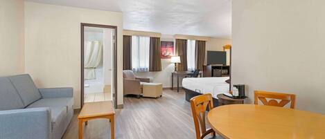 Habitación de hotel con sofá, una mesa de comedor con sillas, cama, escritorio con silla, televisor y baño visible a través de una puerta abierta.