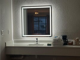 Un baño con lavamanos blanco, un espejo con luz, una ventana y un perchero para toallas.
