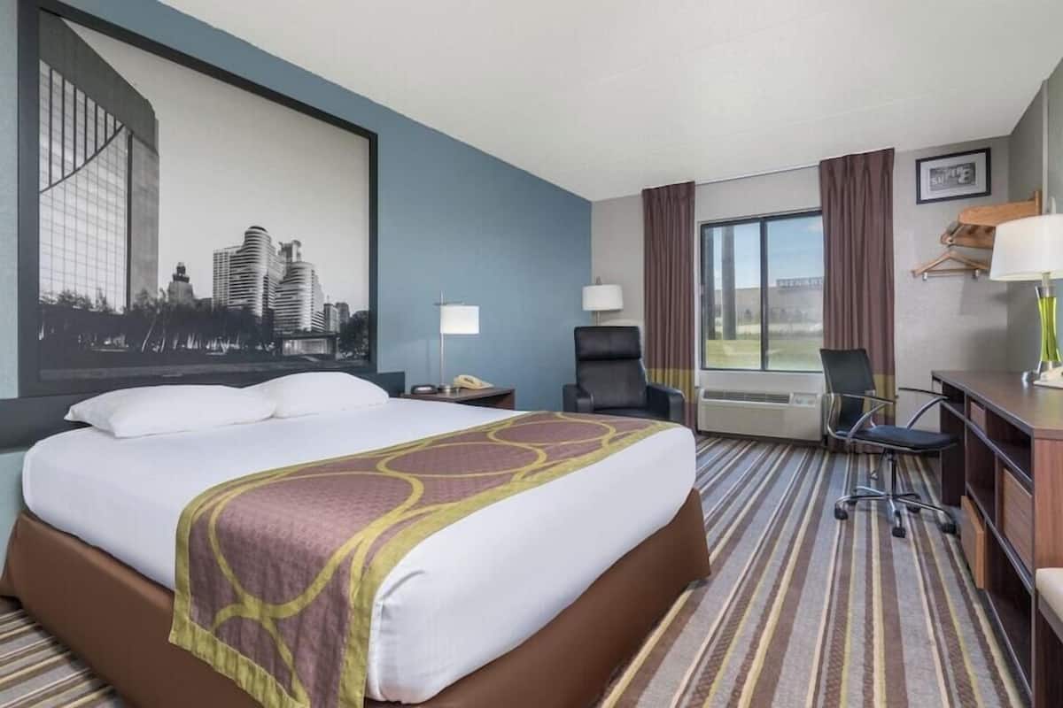 Ein Hotelzimmer mit einem großen Bett, einem Schreibtisch, einem Sessel, einem Fernseher und einem Fenster mit Vorhängen.