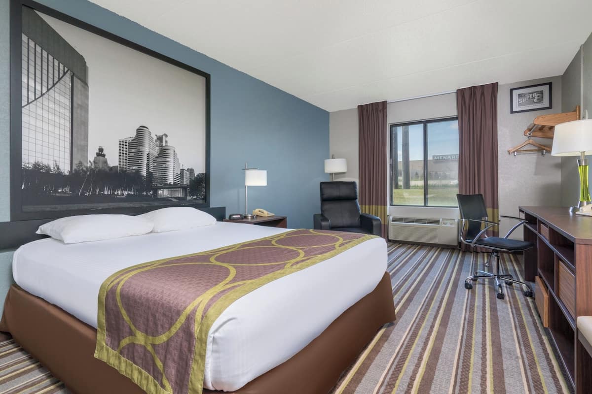 Ein Hotelzimmer mit einem großen Bett, einem Schreibtisch mit Stuhl, einer Schreibtischlampe, einem Fernseher und einem großen Fenster mit Vorhängen.