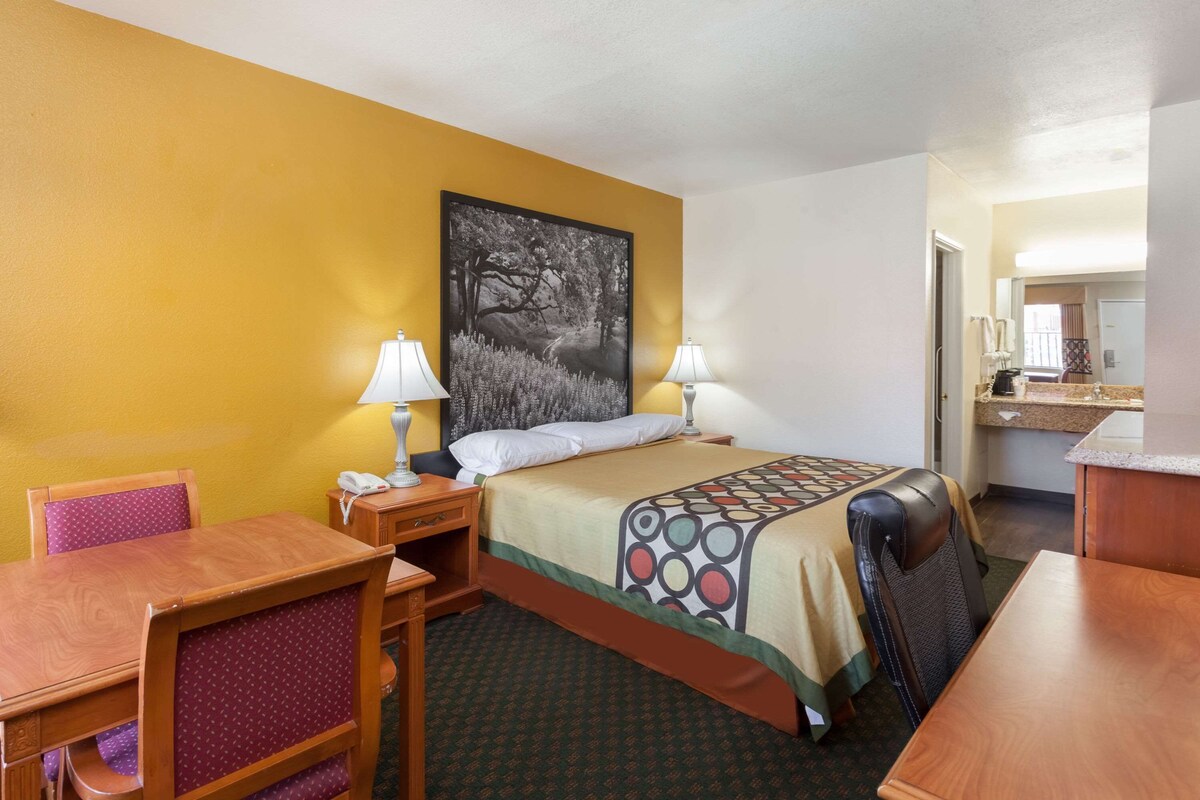A hotel room with a bed, a desk, a chair, two lamps, and a painting on the wall.