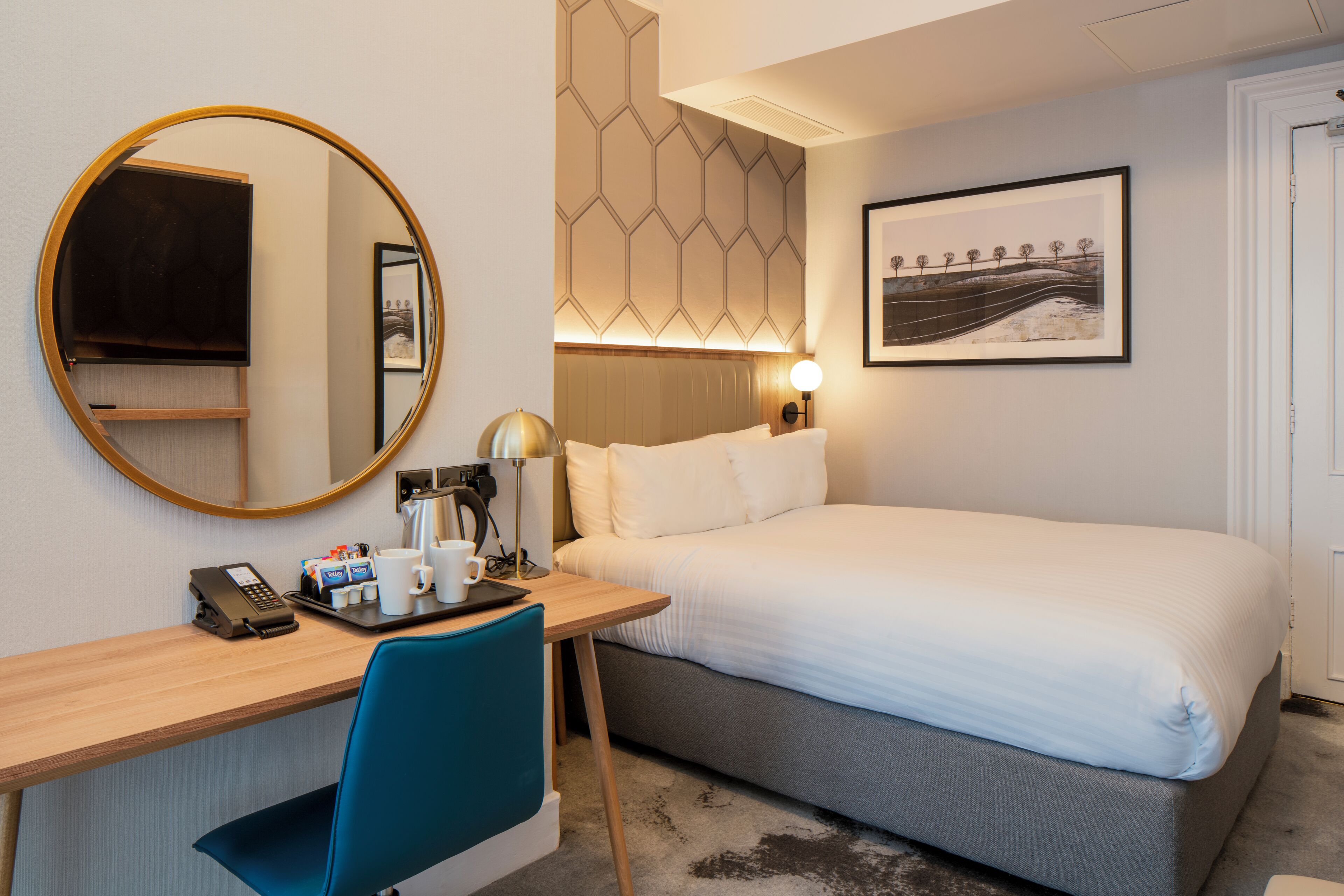 A hotel room with a bed, a desk with a telephone and coffee cups, a chair, a round mirror, a wall-mounted lamp, and a framed picture on the wall.