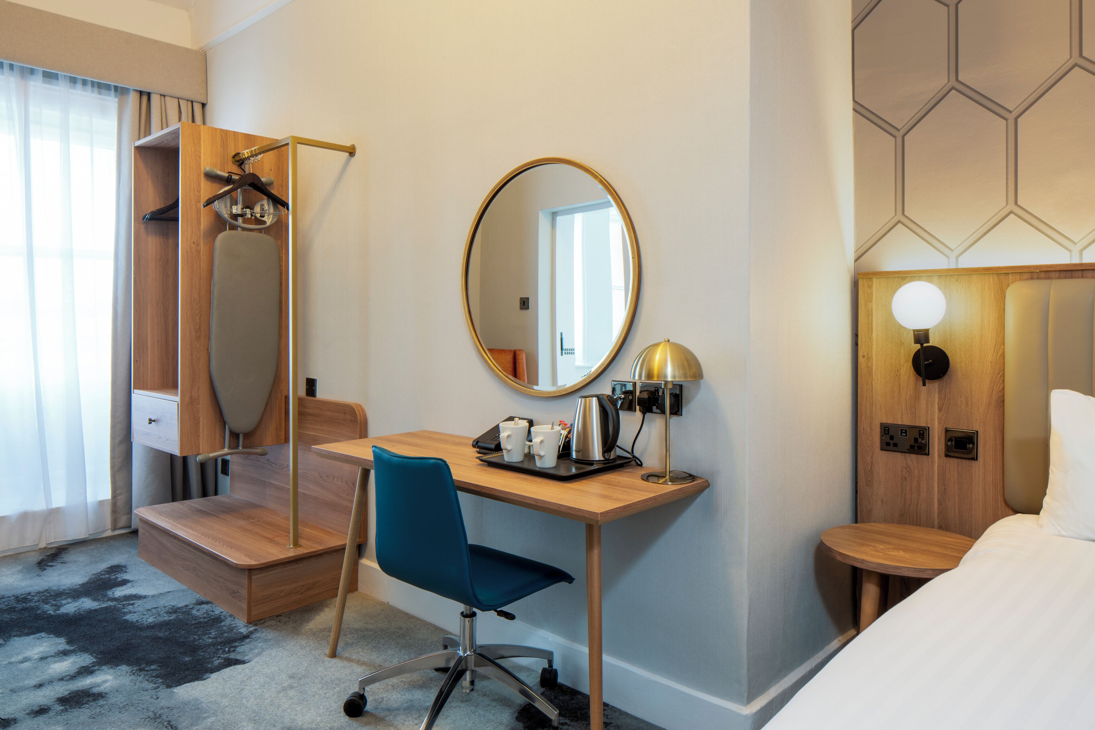 A hotel room with a wooden desk, a blue chair, a mirror, a bedside table with a lamp, and a bed.