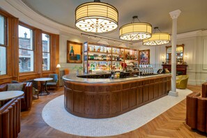 Una zona de bar con mostrador de madera, varias botellas de licor y un área de asientos con sillones de cuero y mesas.