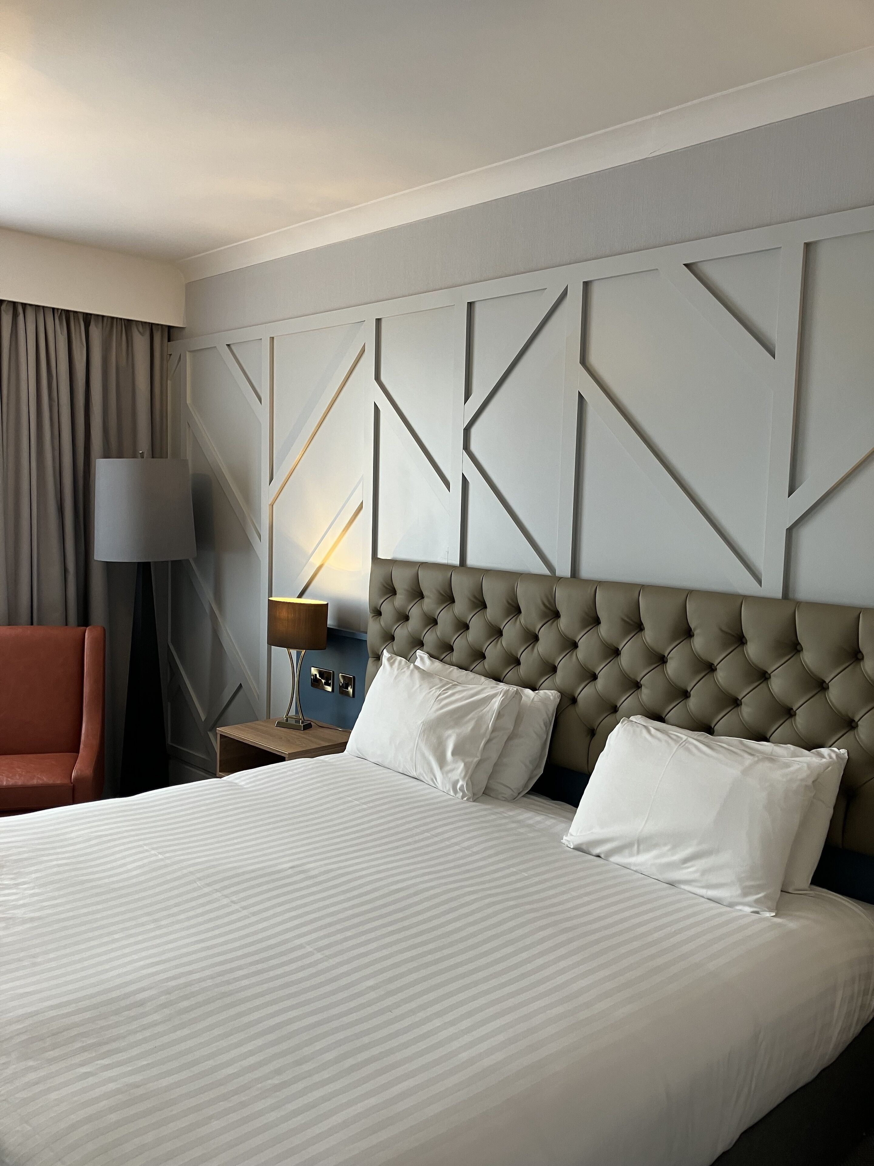 A hotel room with a large bed, a tufted headboard, a nightstand, a lamp, a chair, and a wall with geometric patterns.