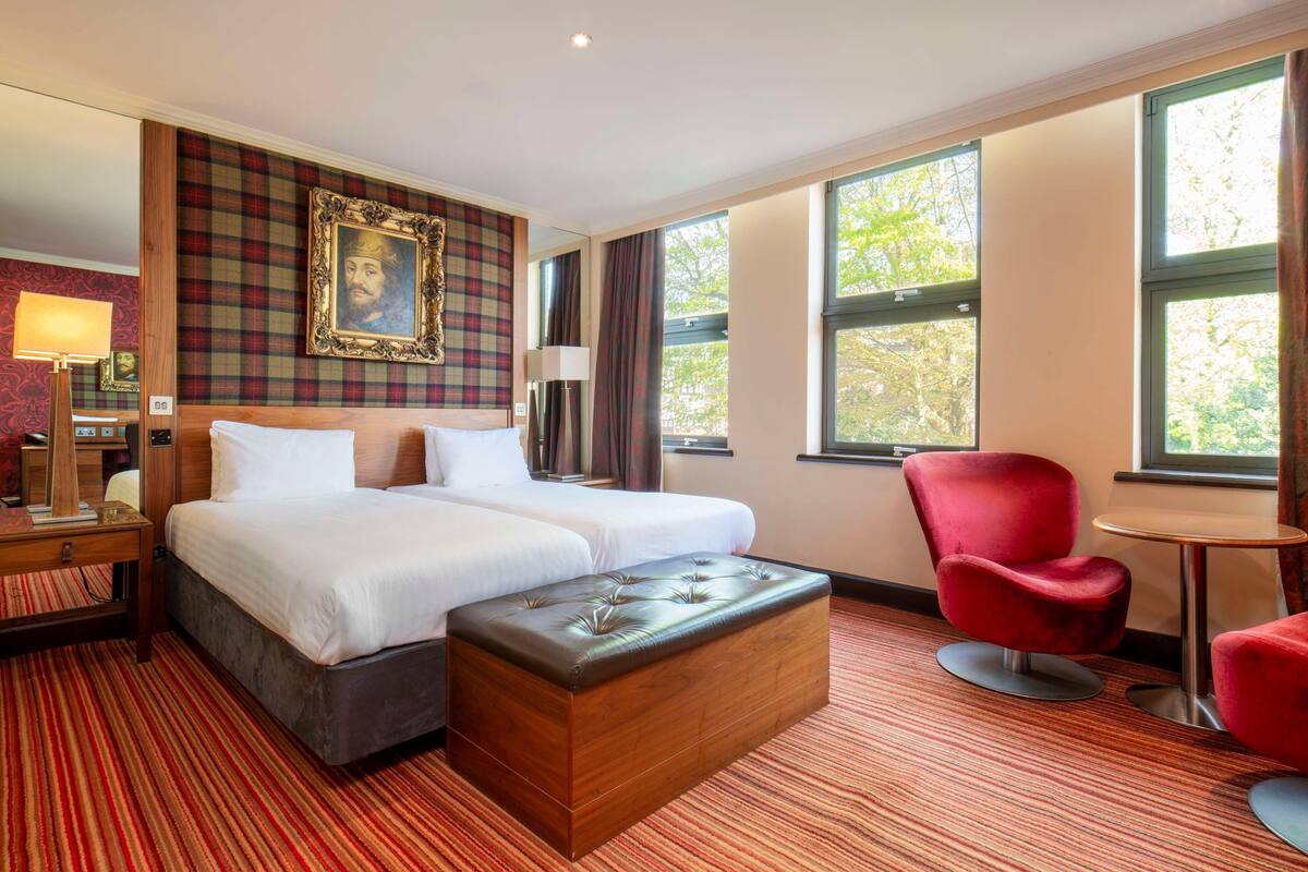 Une chambre d’hôtel avec un grand lit, un fauteuil rouge rembourré, une petite table ronde et une grande fenêtre donnant sur des arbres.