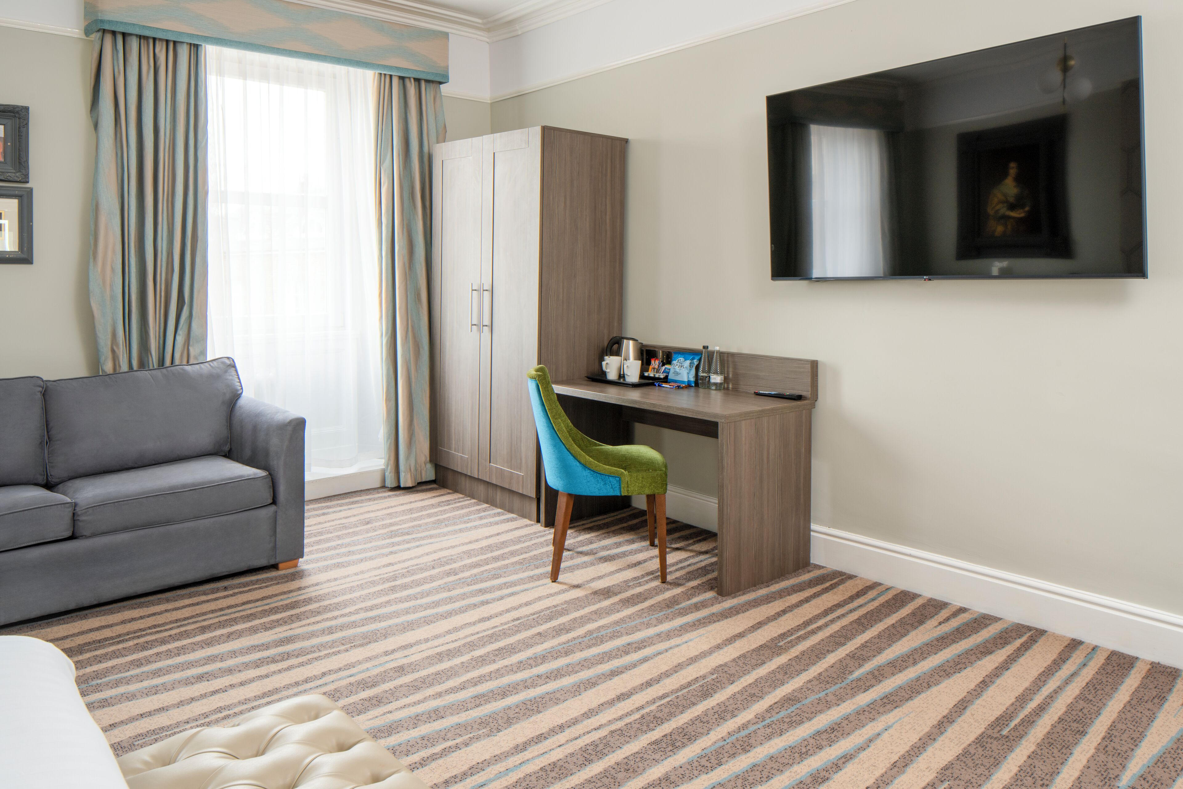 A hotel room with a grey sofa, a desk with a chair, a flat-screen TV, and a patterned carpet.