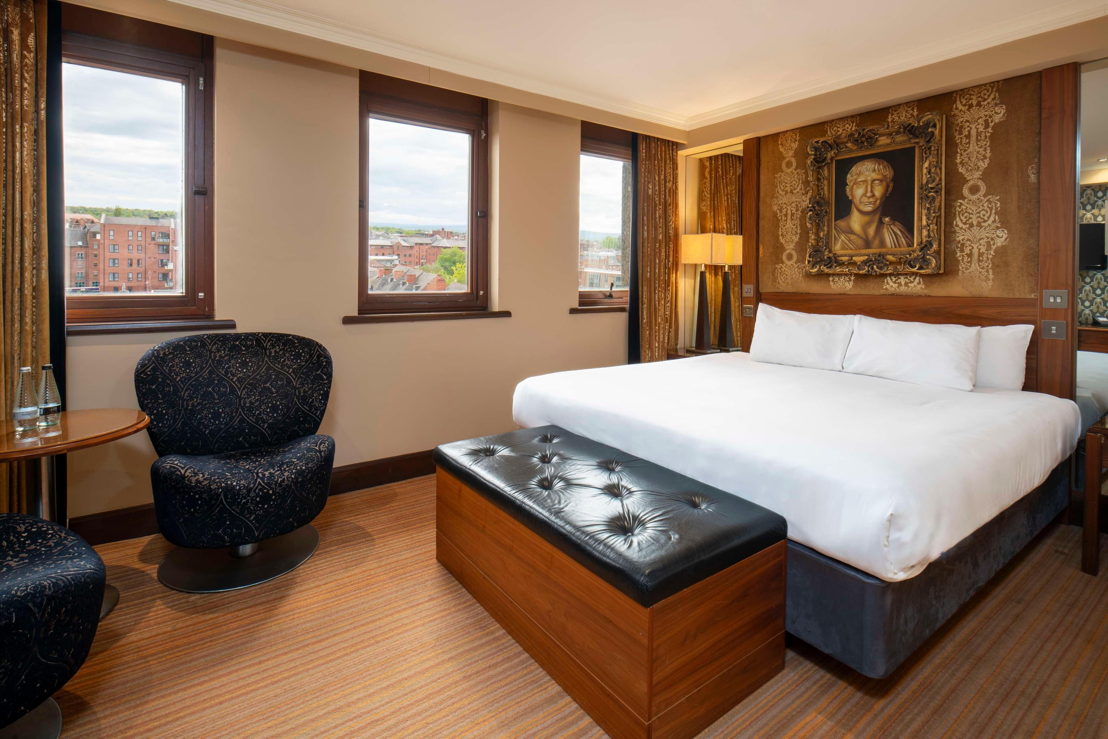 A hotel room with a large bed, a wooden chest of drawers, a round table, a blue upholstered chair, and a large window with a city view.
