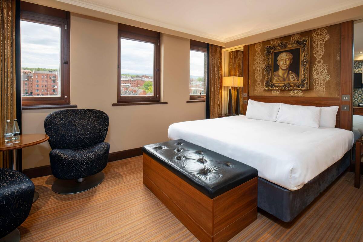 A hotel room with a large bed, a wooden chest of drawers, a round table, a blue upholstered chair, and a large window with a city view.