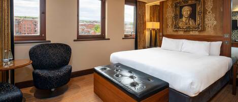 A hotel room with a large bed, a wooden chest of drawers, a round table, a blue upholstered chair, and a large window with a city view.