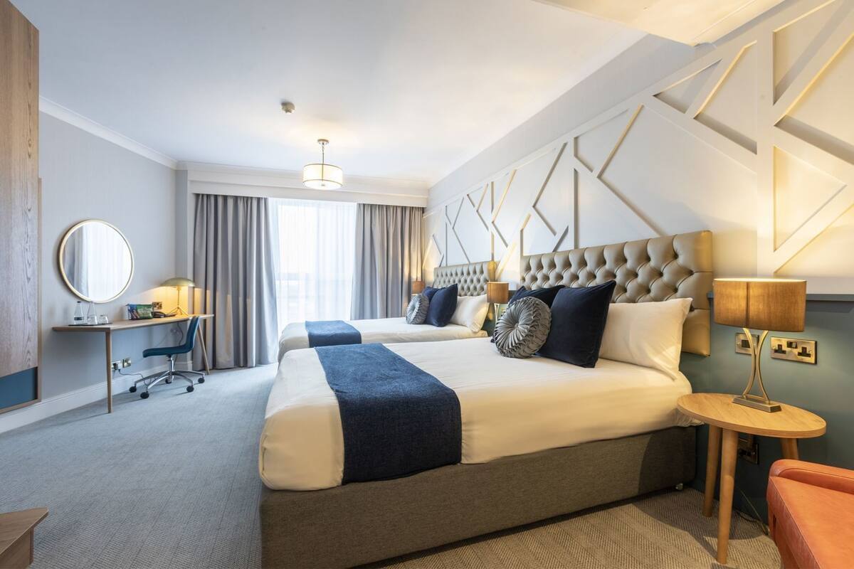 Habitación de hotel con dos camas, un escritorio, una silla, un espejo redondo y una lámpara.