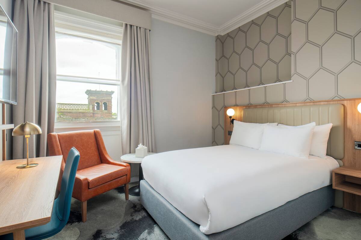 A hotel room with a large bed, a desk with a chair, a small table, and a view of a building through the window.