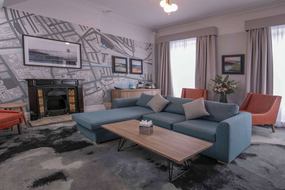 Un salon comprenant un canapé d’angle, deux fauteuils, une table basse, une cheminée et un mur décoré d’une carte géographique.