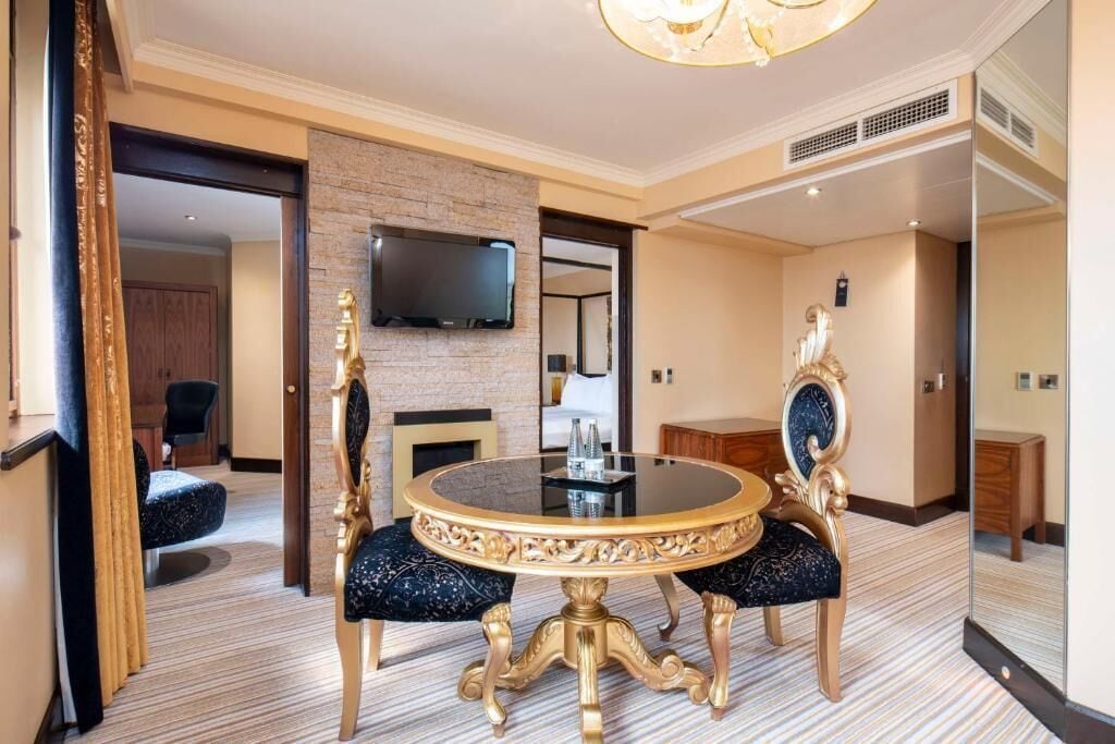Habitación de hotel con una mesa de comedor redonda, dos sillas ornamentadas, un televisor de pantalla plana montado en una pared de ladrillo y un espejo grande.