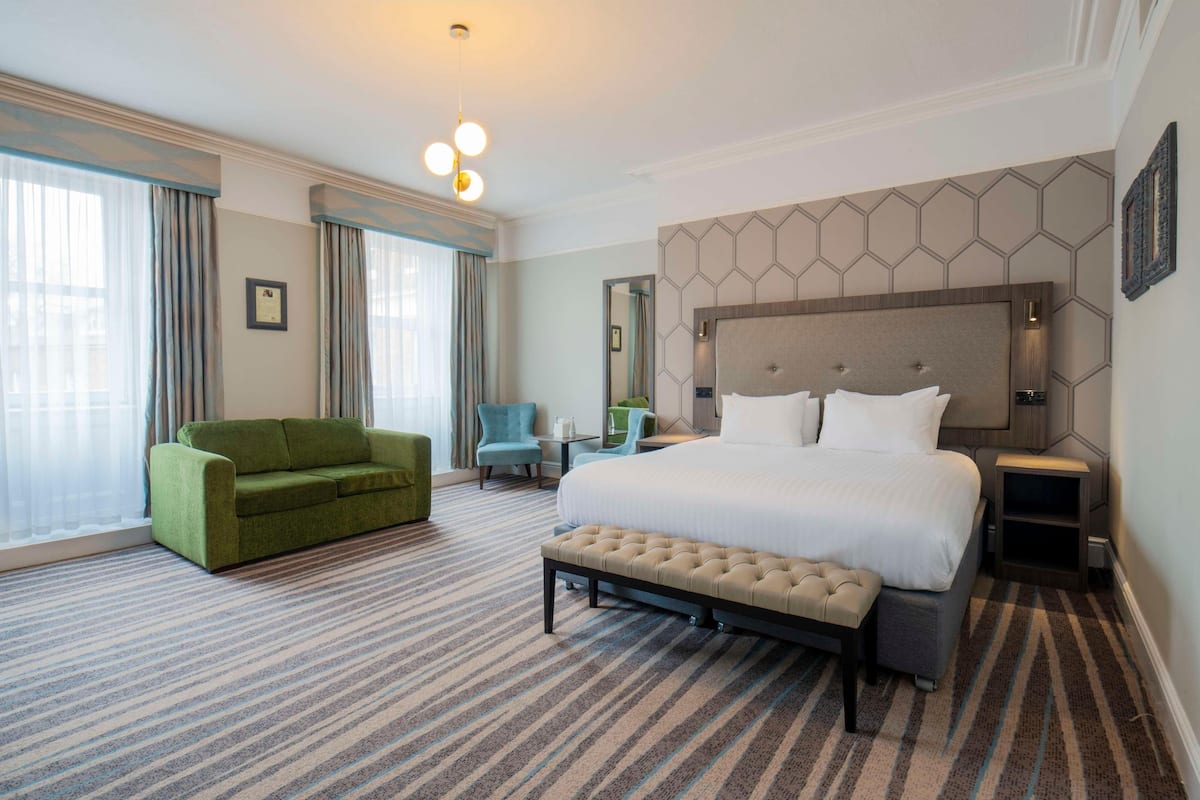 A hotel room with a large bed, a green sofa, a small table, and a chair.