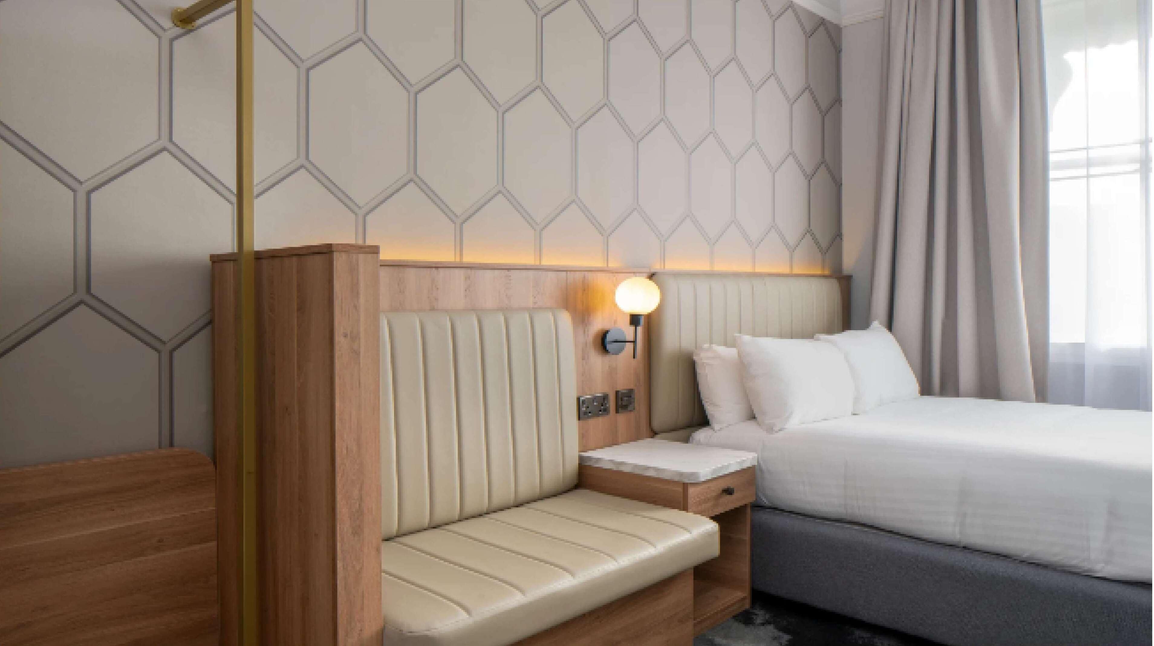 A hotel room with a bed, a bedside table, a lamp, a bench, and a wall with a geometric pattern.