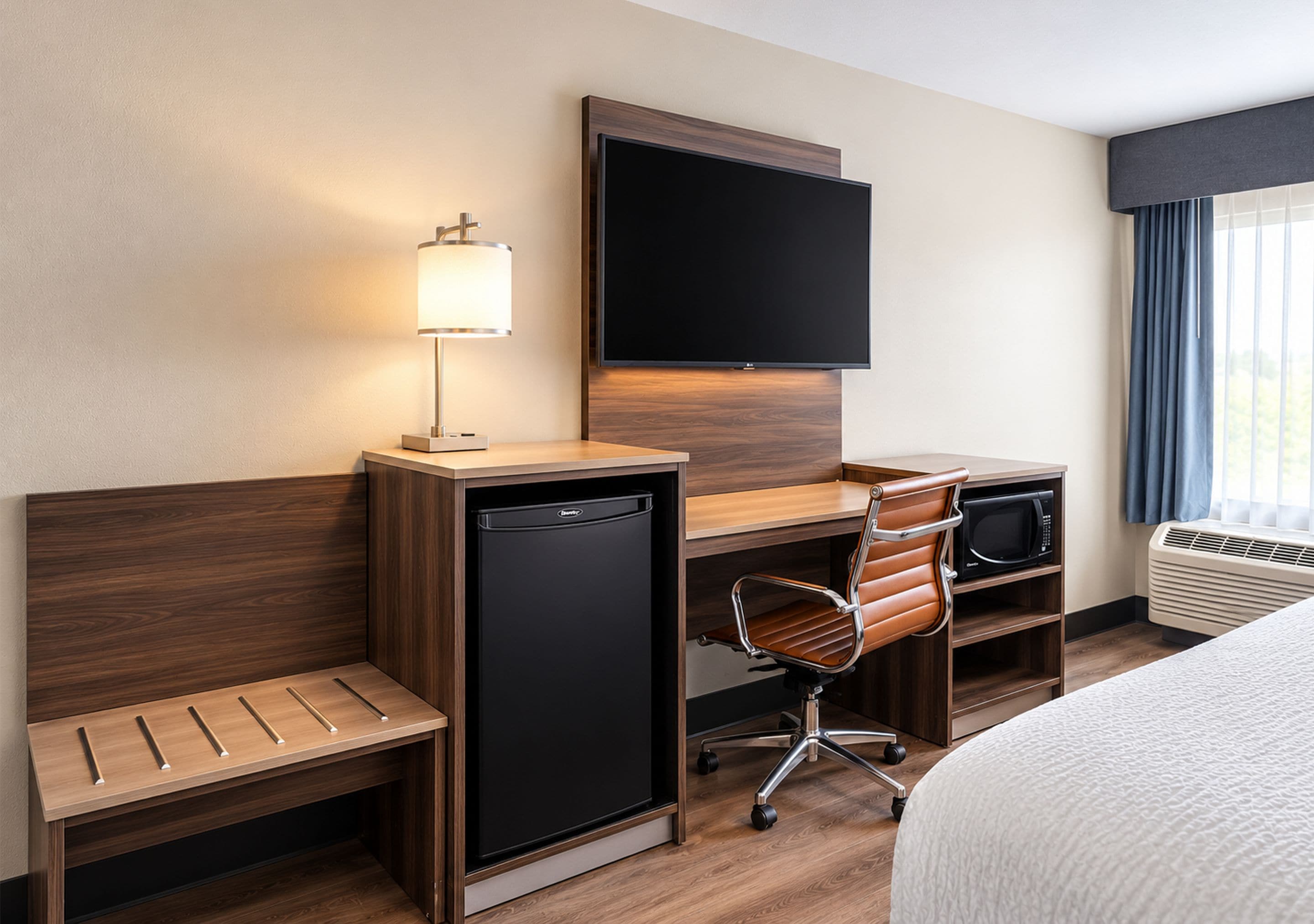 A hotel room with a bed, a desk with a computer, a chair, a small refrigerator, a TV, and a window with curtains.