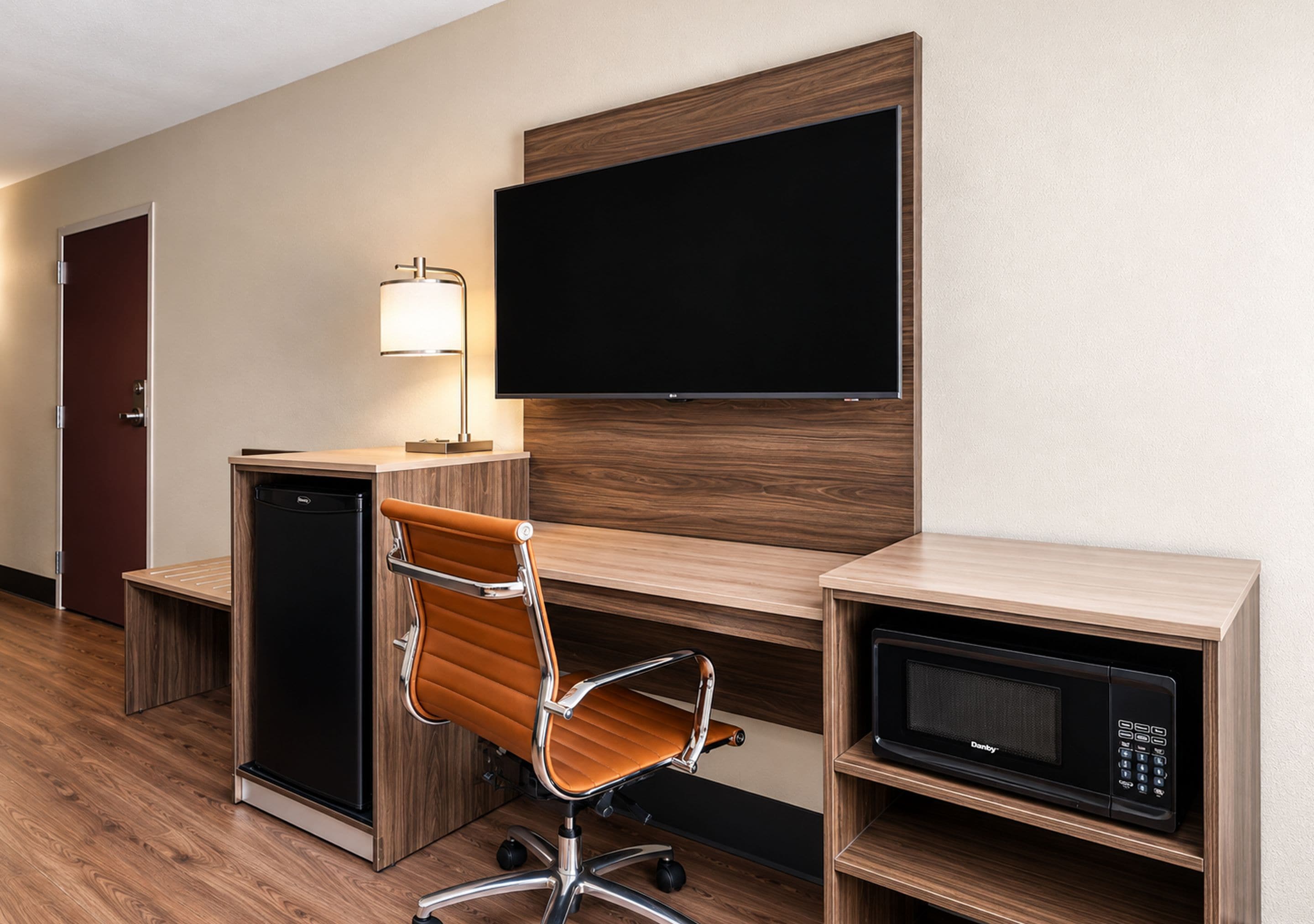 A hotel room with a desk, a chair, a television, a lamp, a microwave, and a mini-fridge.