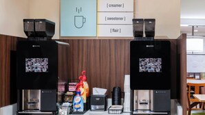 Una estación de café con dos máquinas de café, un cartel que indica la adición de leche condensada, endulzantes y aromas, además de un dispensador de café.