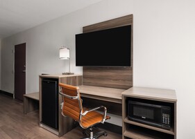 Habitación de hotel con escritorio de madera, una silla moderna, un televisor de pantalla plana, una lámpara, microondas y un mini refrigerador.