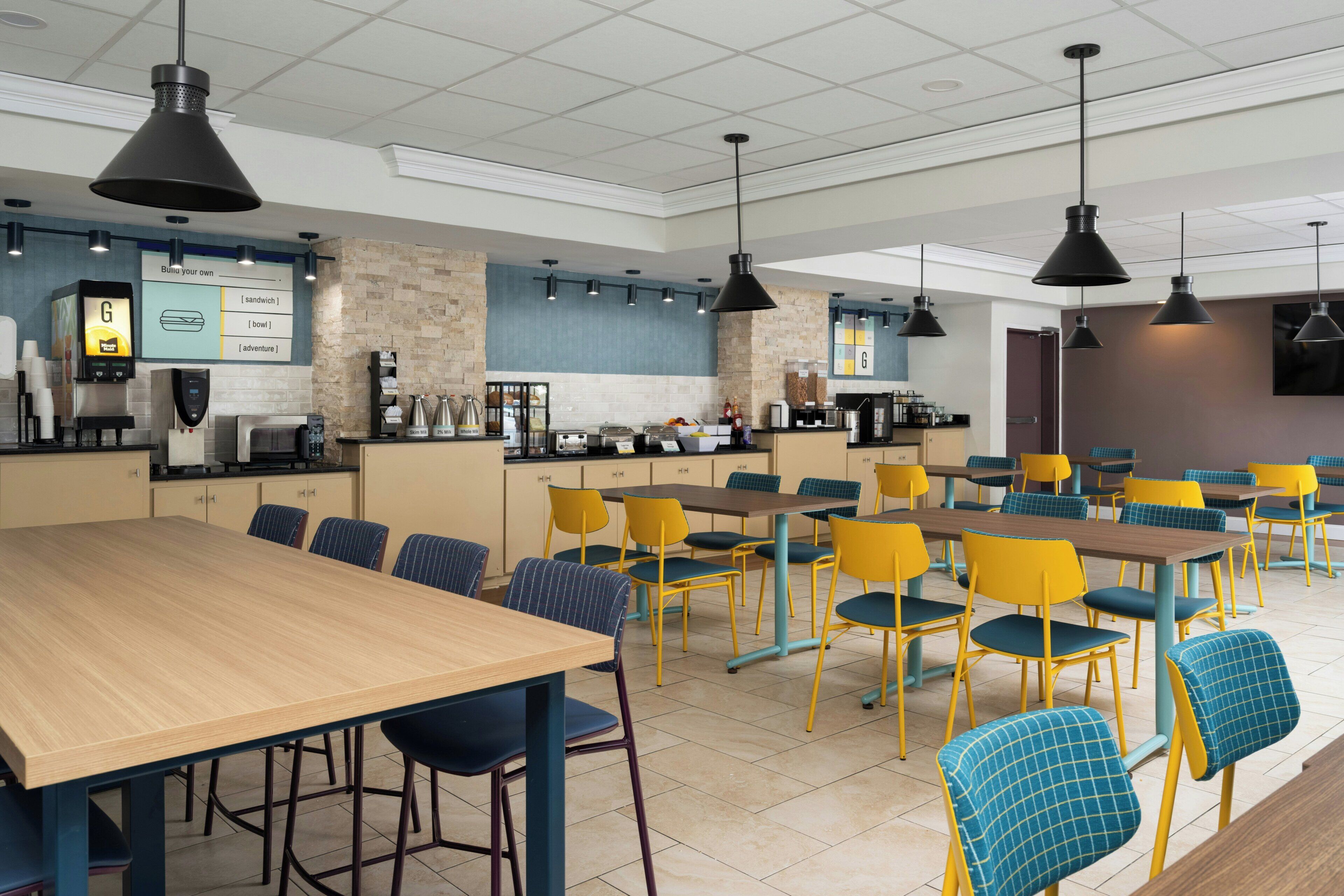 Une cafétéria moderne avec de longues tables en bois et des chaises jaunes, équipée de machines à café et offrant un large choix de plats.