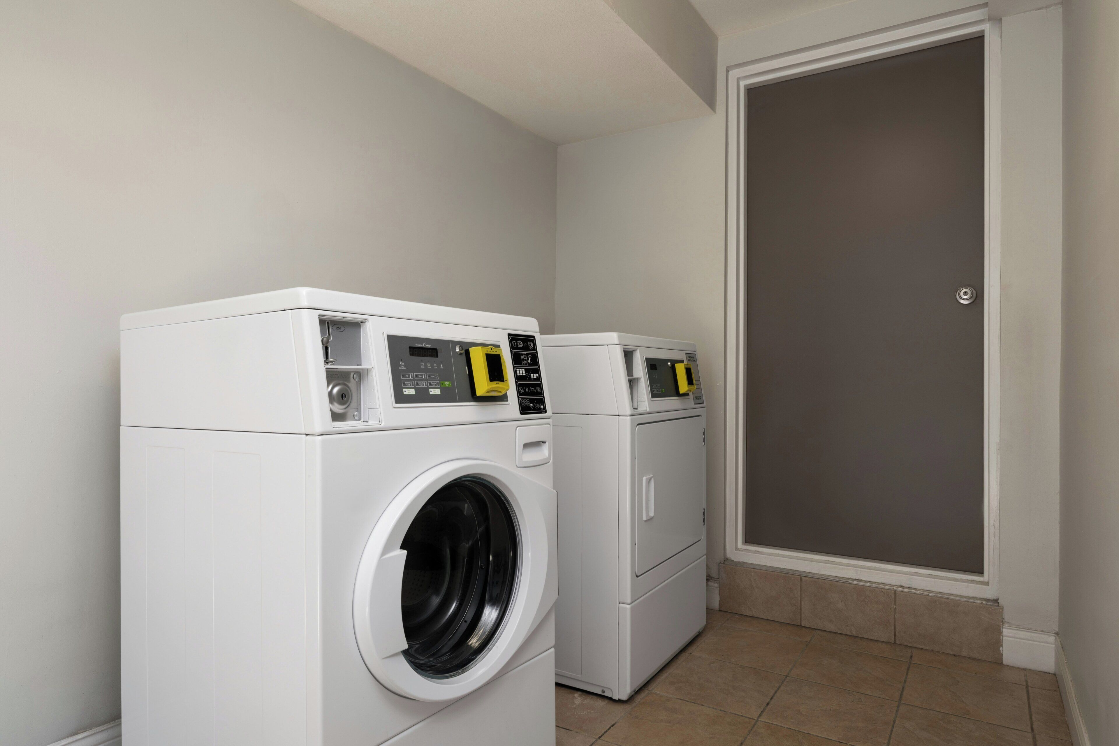 A laundry room with two white washing machines and a dryer, a tiled floor, and a door.