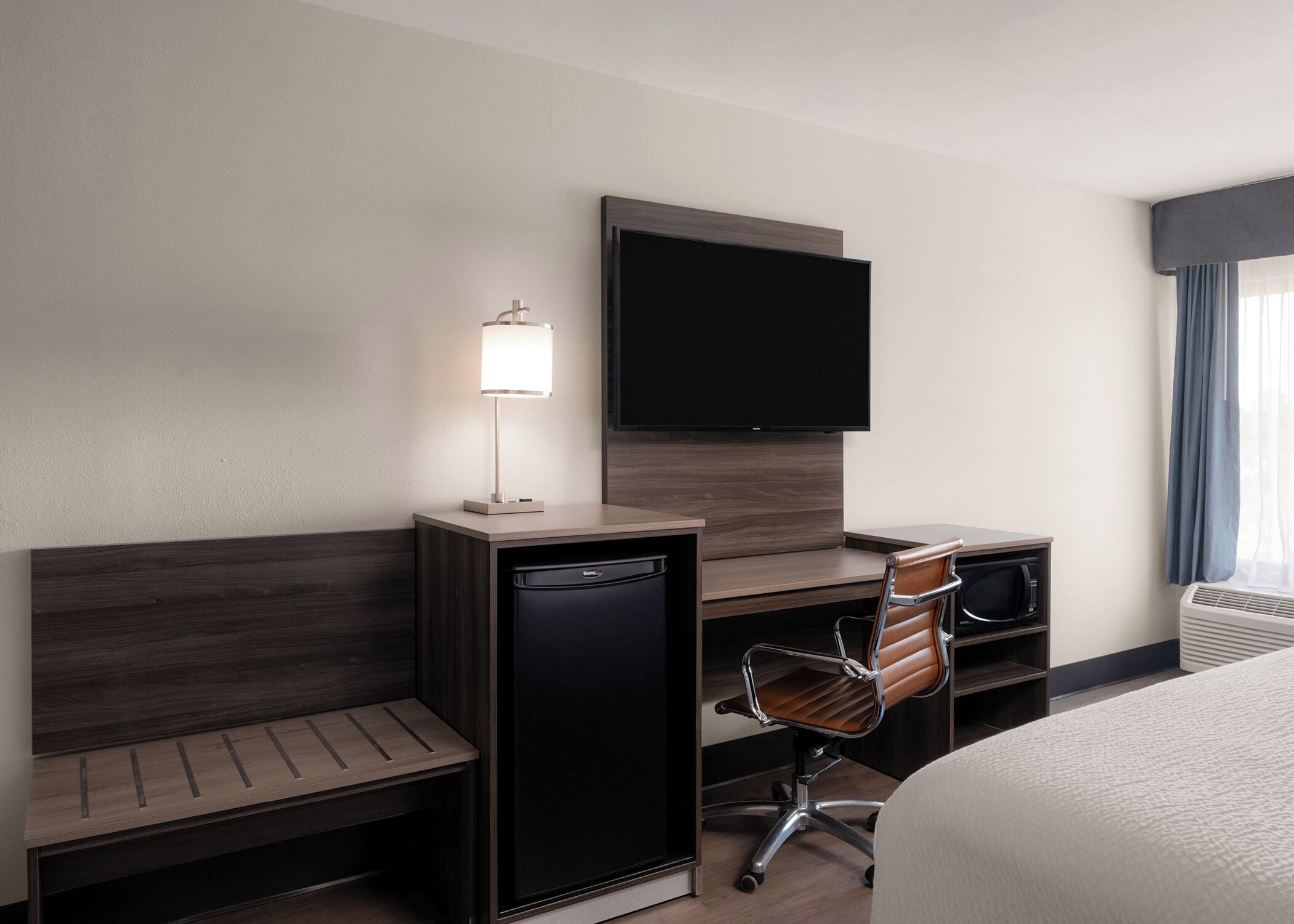 Camera d'albergo con un letto, una scrivania con televisore a schermo piatto, una lampada, una sedia, un forno a microonde e un mini-frigorifero.