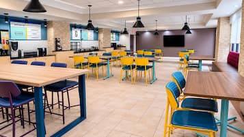 A modern cafeteria with multiple tables and chairs, a counter with a menu board, and a television screen.
