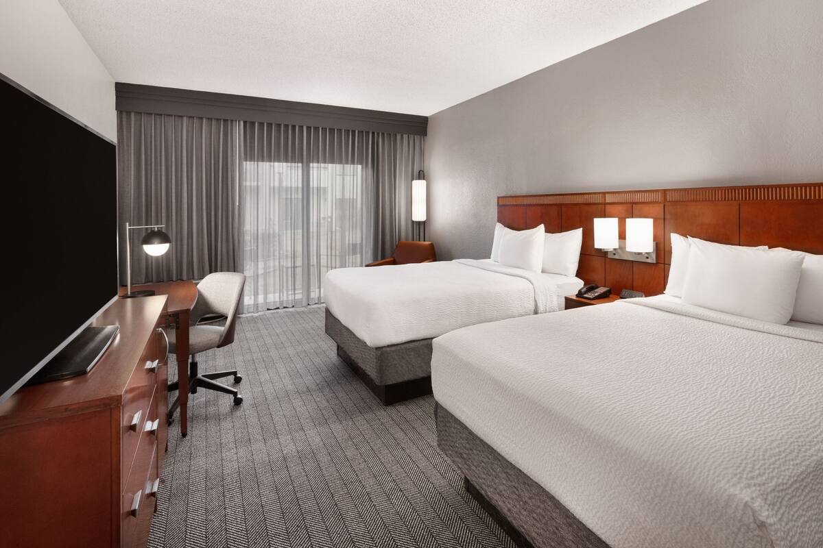 A hotel room with two beds, a desk, a chair, a TV, and a large window with curtains.