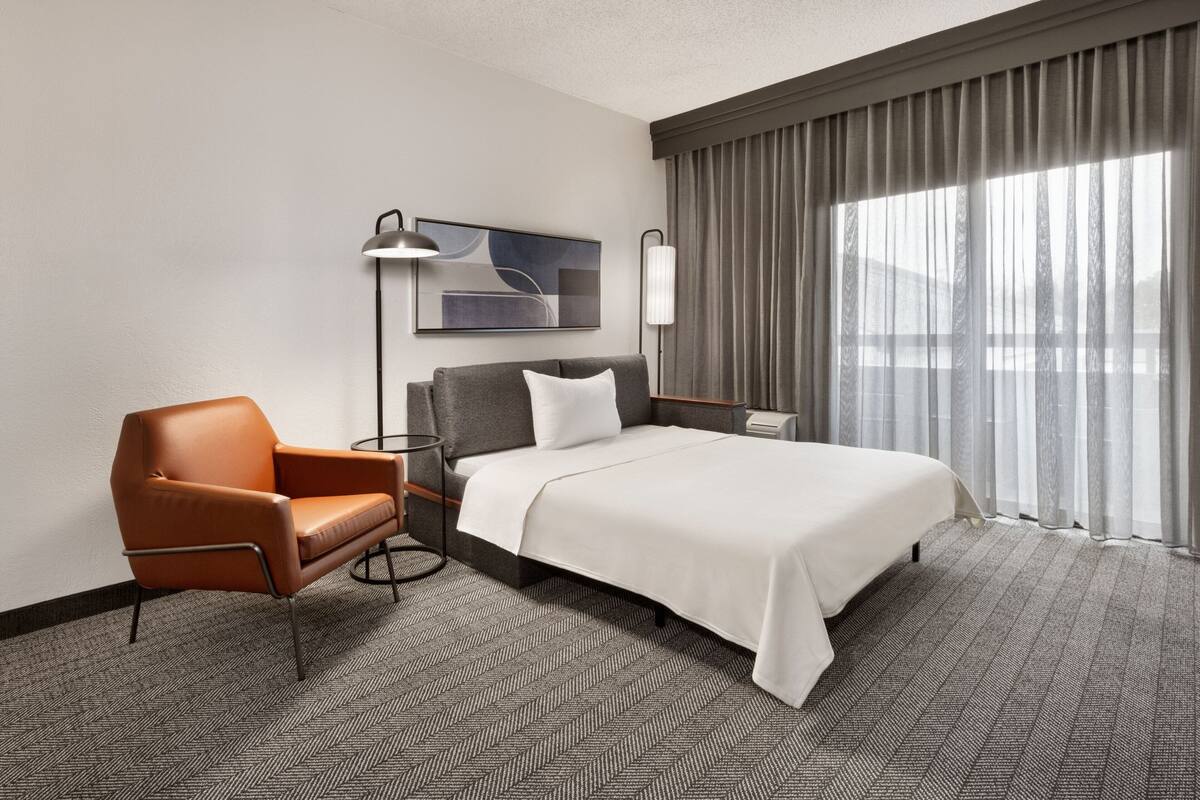 A hotel room with a bed, a chair, a lamp, and a painting on the wall.