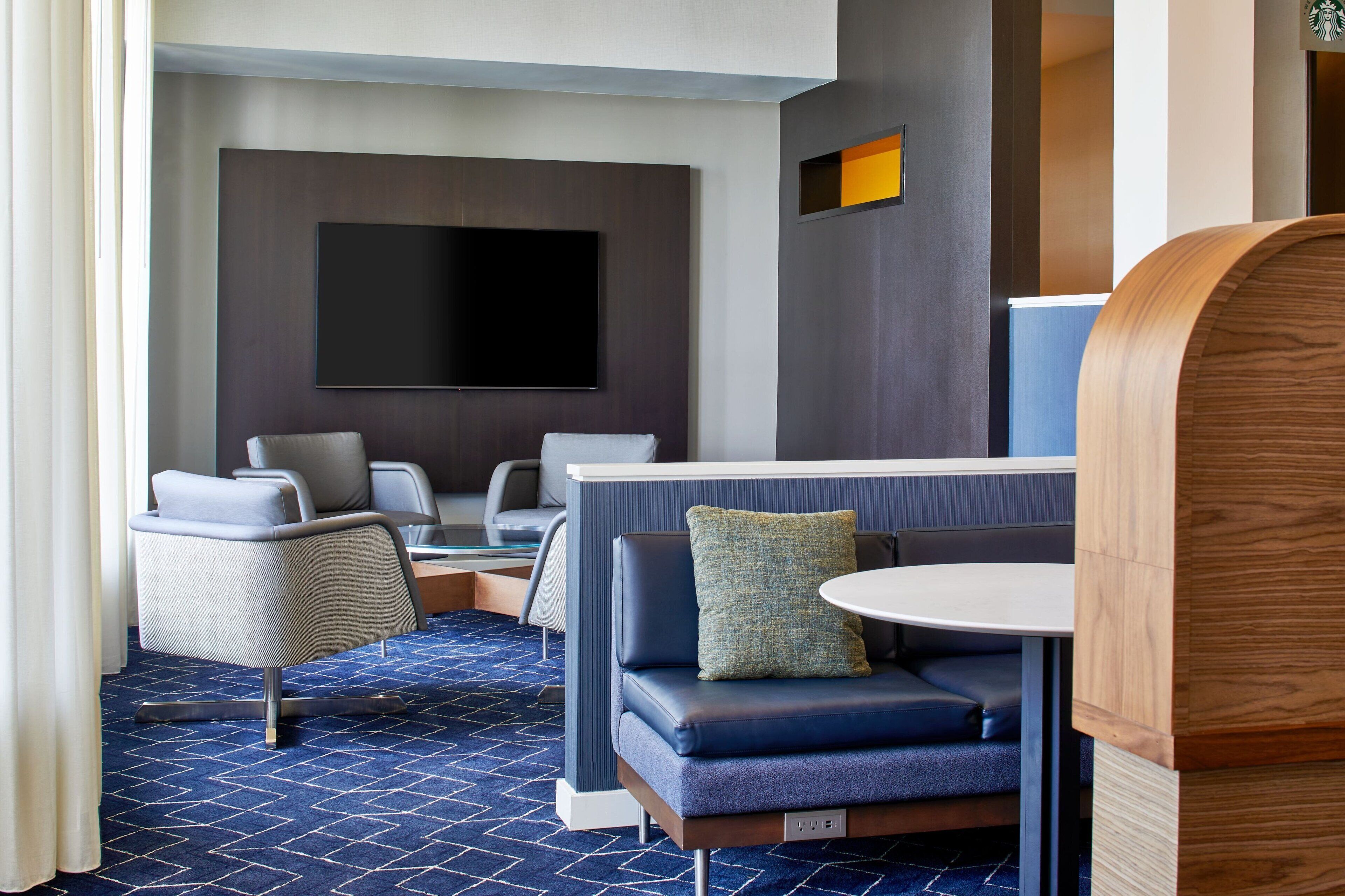 A modern hotel room with a large flat-screen TV mounted on the wall, a seating area with a sofa and chairs, and a small round table.