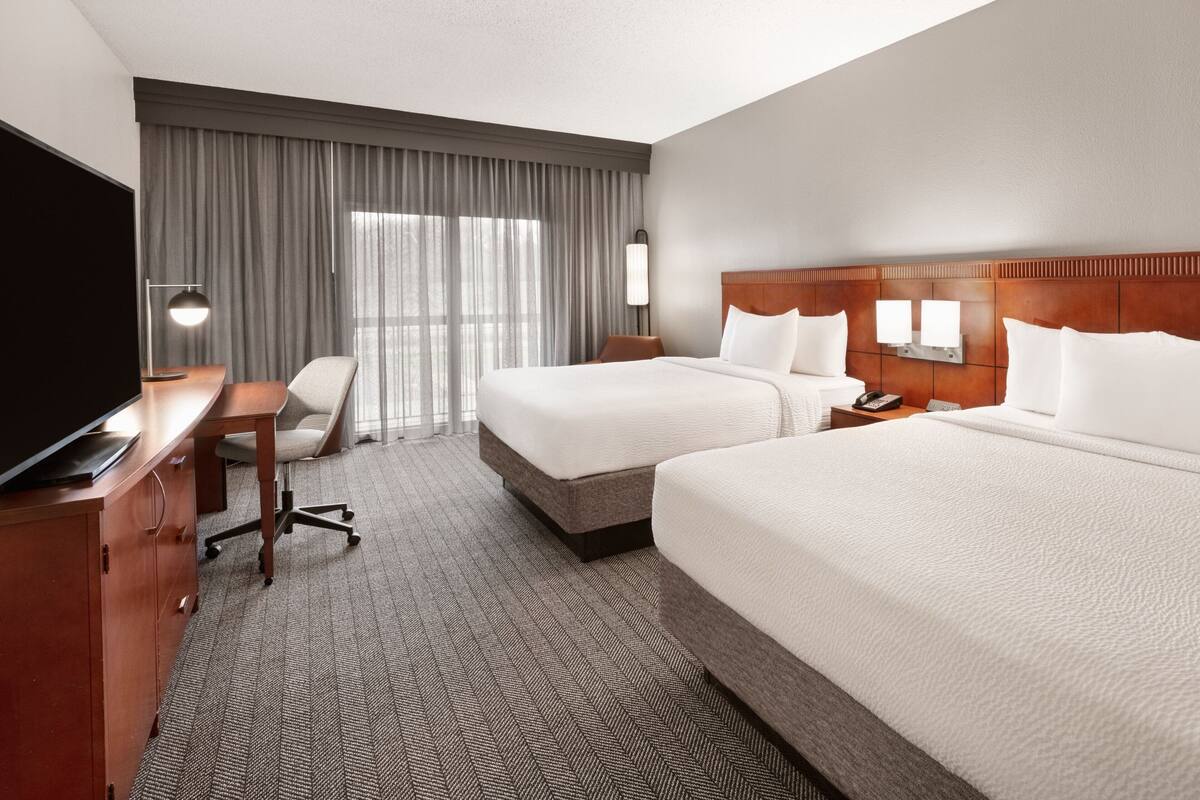 A hotel room with two beds, a desk, a chair, a TV, and a large window with curtains.