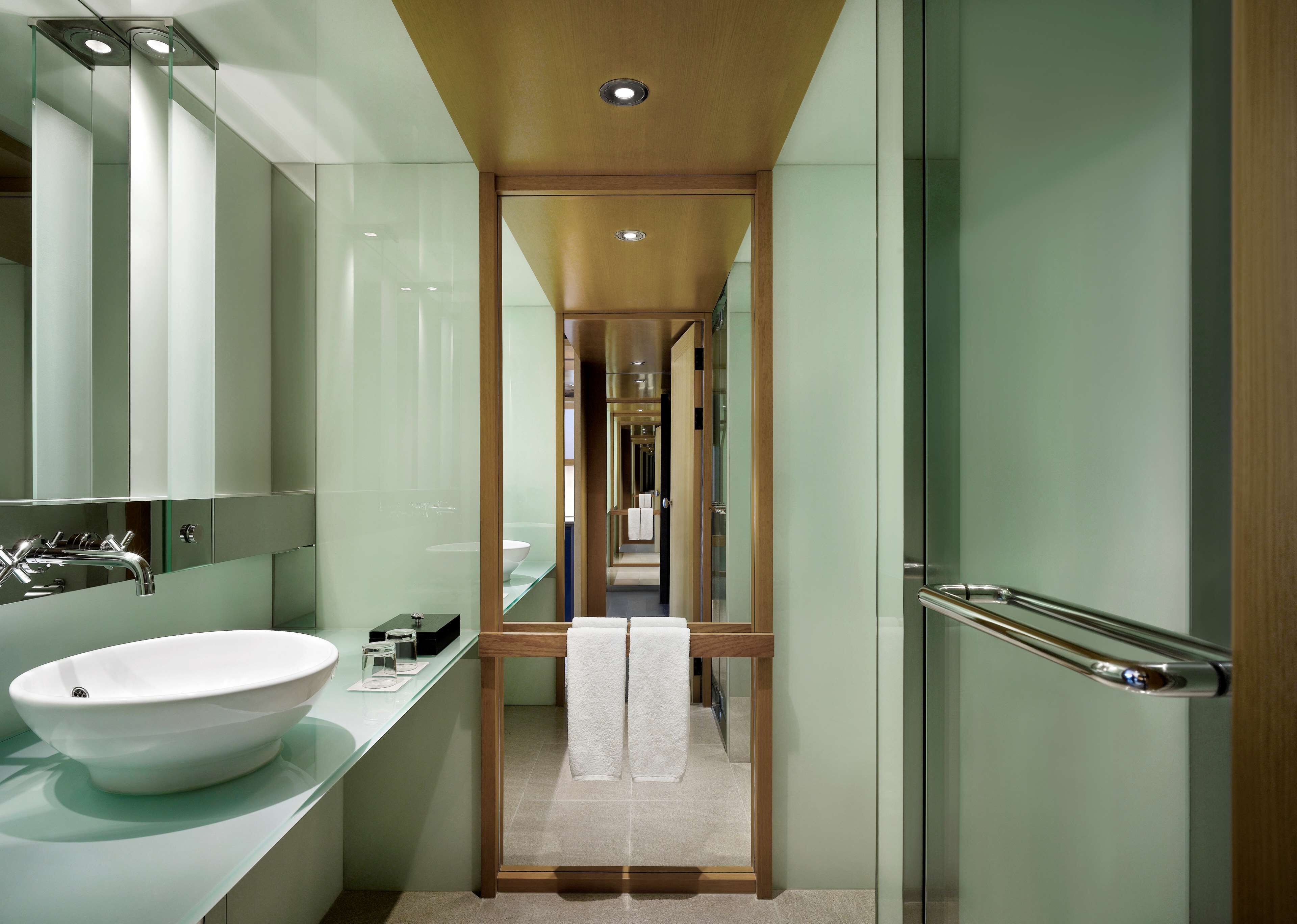 Un baño moderno con un lavamanos grande de color blanco, una encimera de vidrio y un espejo encima. Se ve una puerta de madera que da a un pasillo.