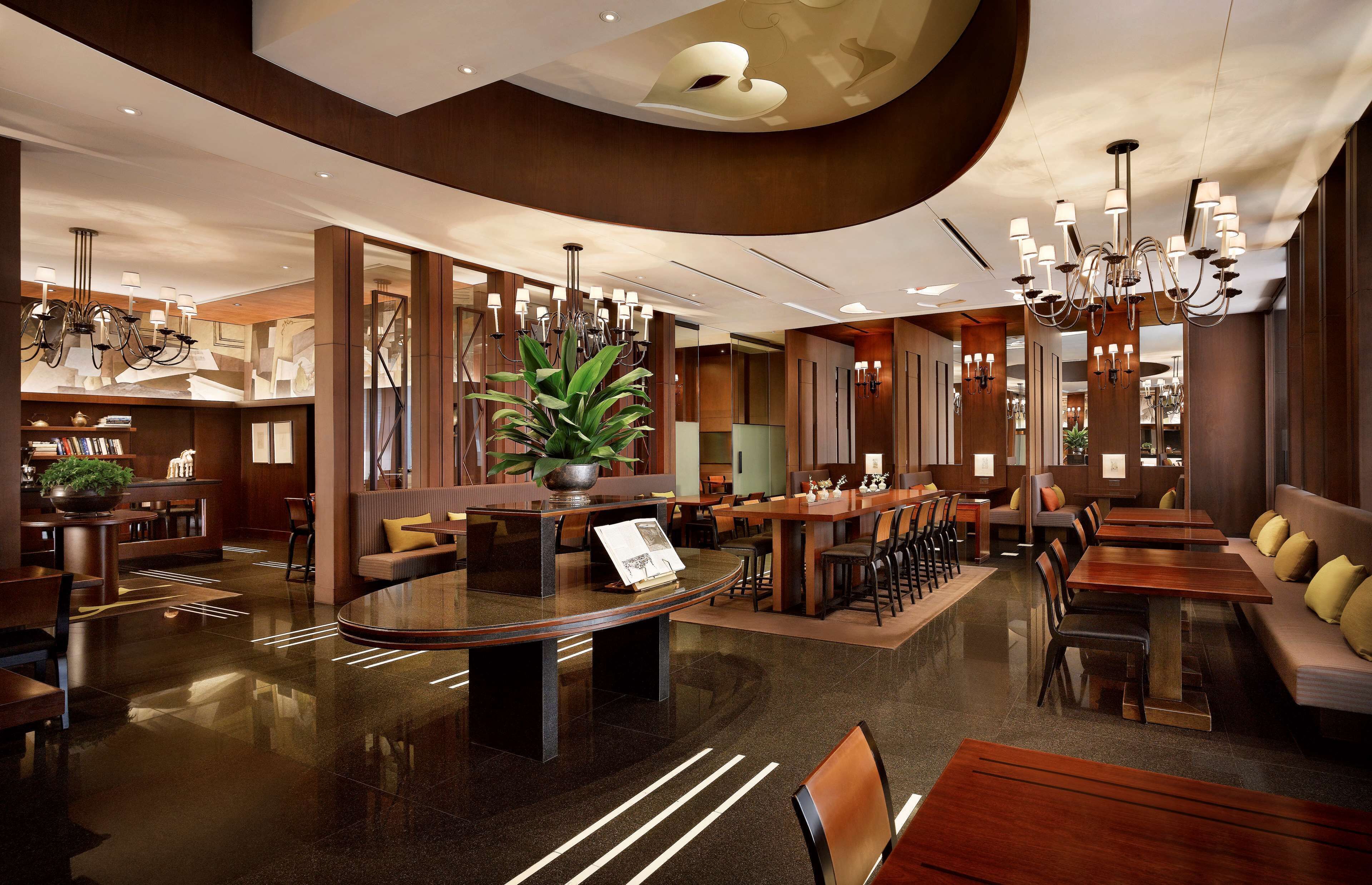 A modern hotel lobby with a central round table, seating areas, and a large chandelier.
