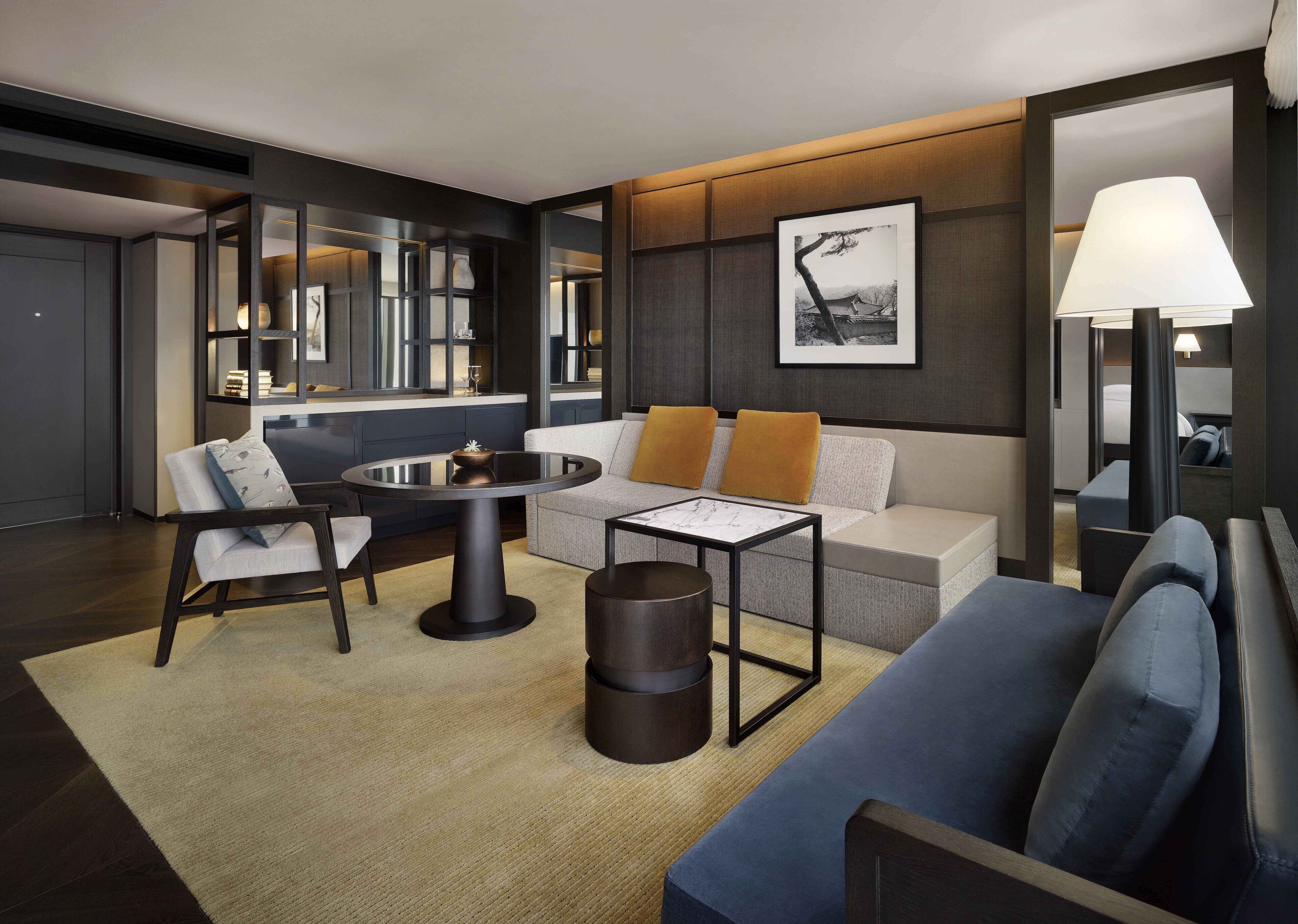 Una habitación de hotel moderna con una mesa redonda de comedor, un sofá con cojines naranja, una mesita auxiliar y un espejo grande que refleja la habitación.