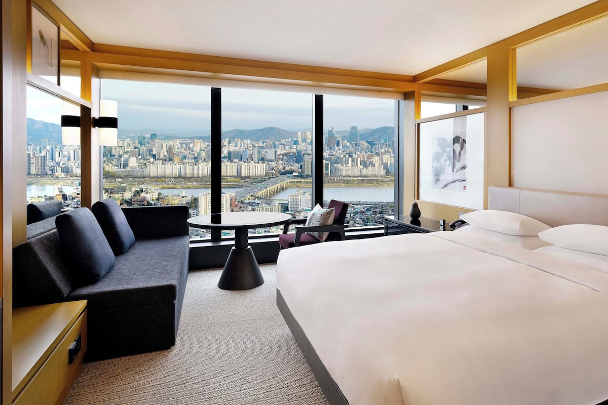 A modern hotel room with a large bed, a sofa, a small table, and a view of a cityscape through large windows.