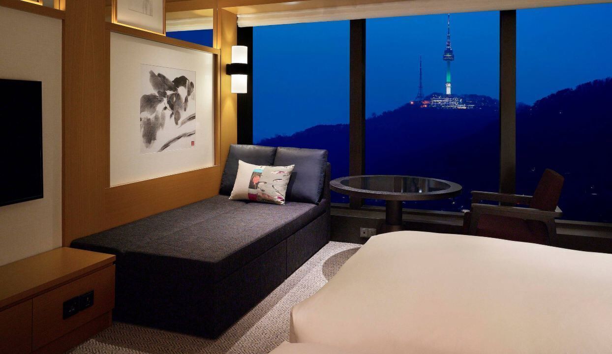 A hotel room with a bed, a small seating area, a table, and a large window with a view of a tower.