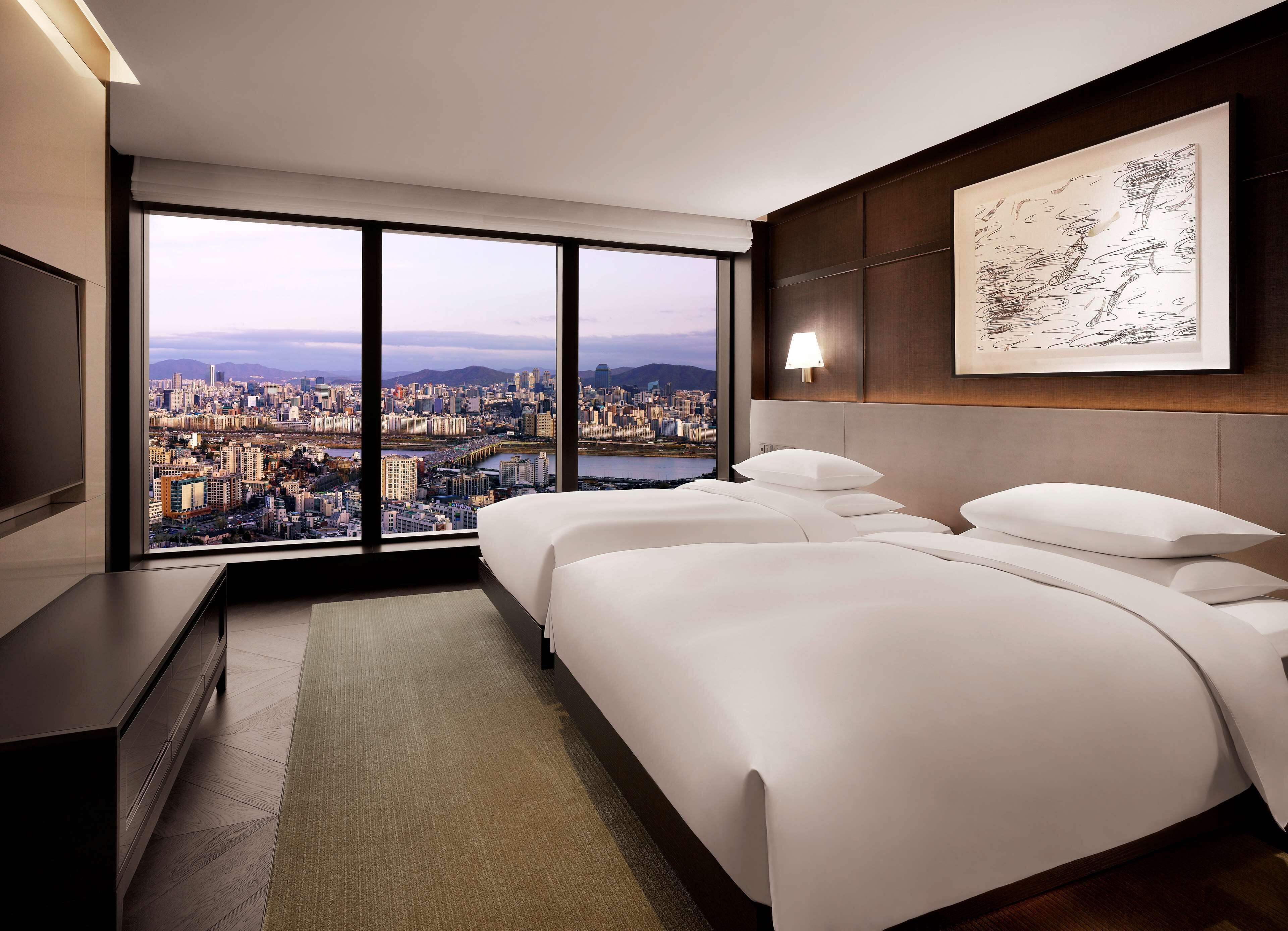 A hotel room with a large window offering a city view, a bed with white linens, a bedside table, and a wall-mounted artwork.