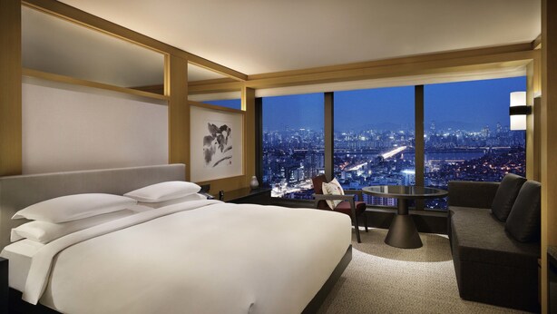 Habitación de hotel con una cama grande, una mesa de comedor y un sofá. Se ve la ciudad a través de amplios ventanales.