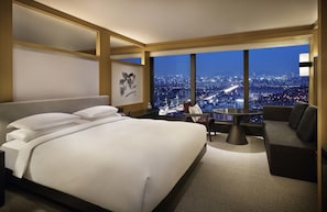 Habitación de hotel con una cama grande, una mesa de comedor y un sofá. Se ve la ciudad a través de amplios ventanales.