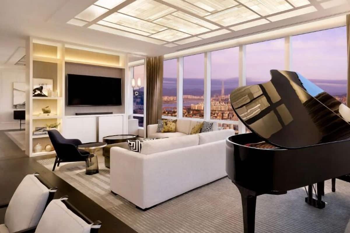 A modern living room with a grand piano, a large flat-screen TV, and a city view through large windows.