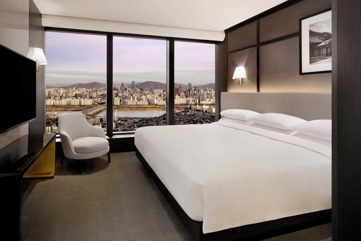 A hotel room with a large bed, a chair, a TV, and a city view through large windows.