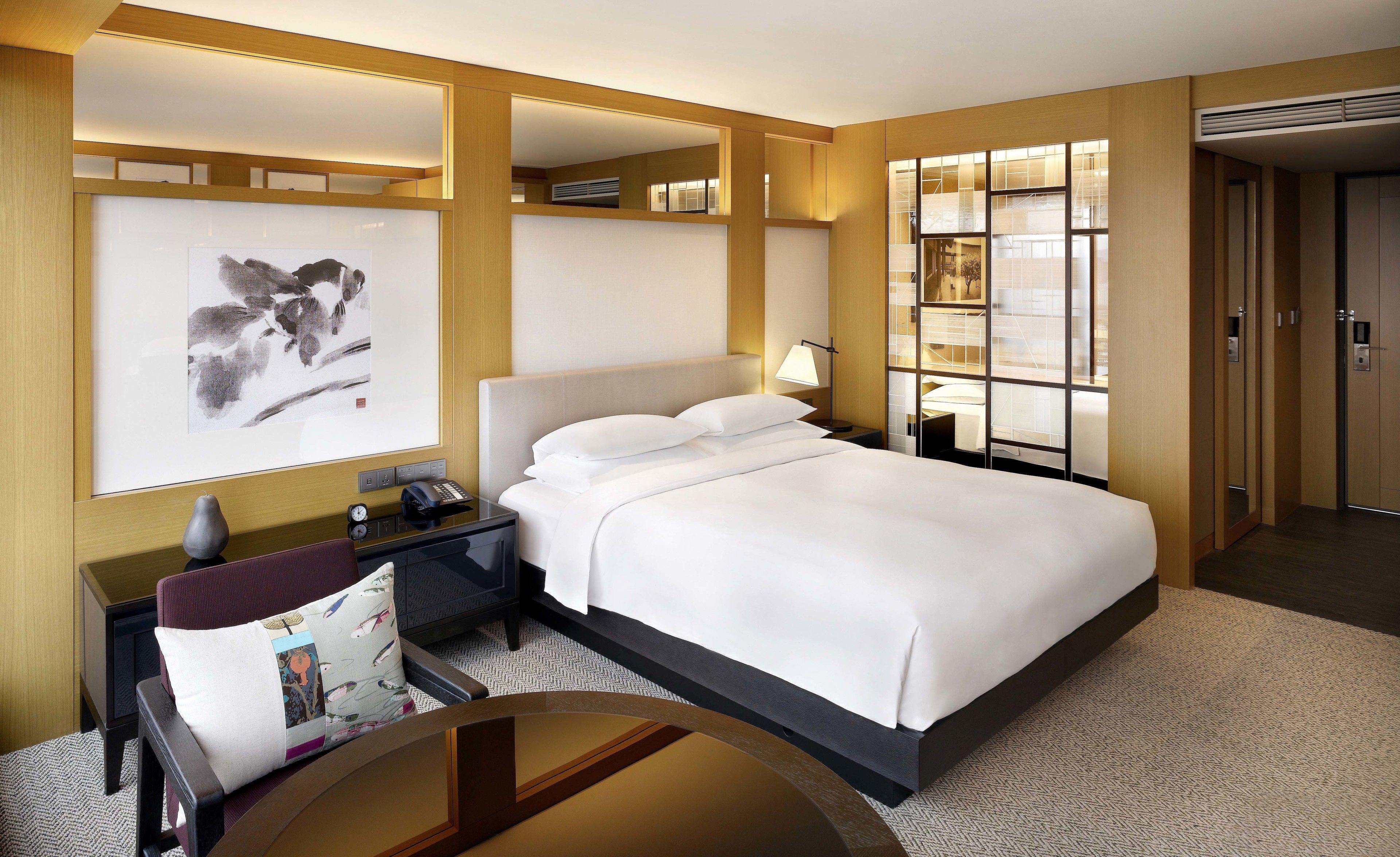 Habitación de hotel con una cama grande, un escritorio con lámpara, una obra de arte en la pared y vistas de la ciudad a través de una ventana.