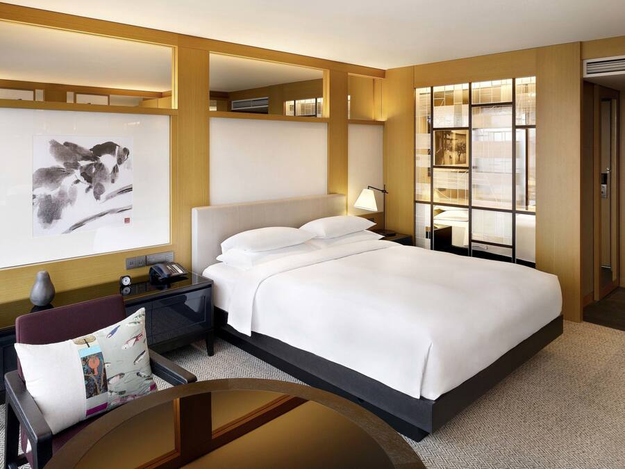 Habitación de hotel con una cama grande, un escritorio con lámpara, una obra de arte en la pared y vistas de la ciudad a través de una ventana.