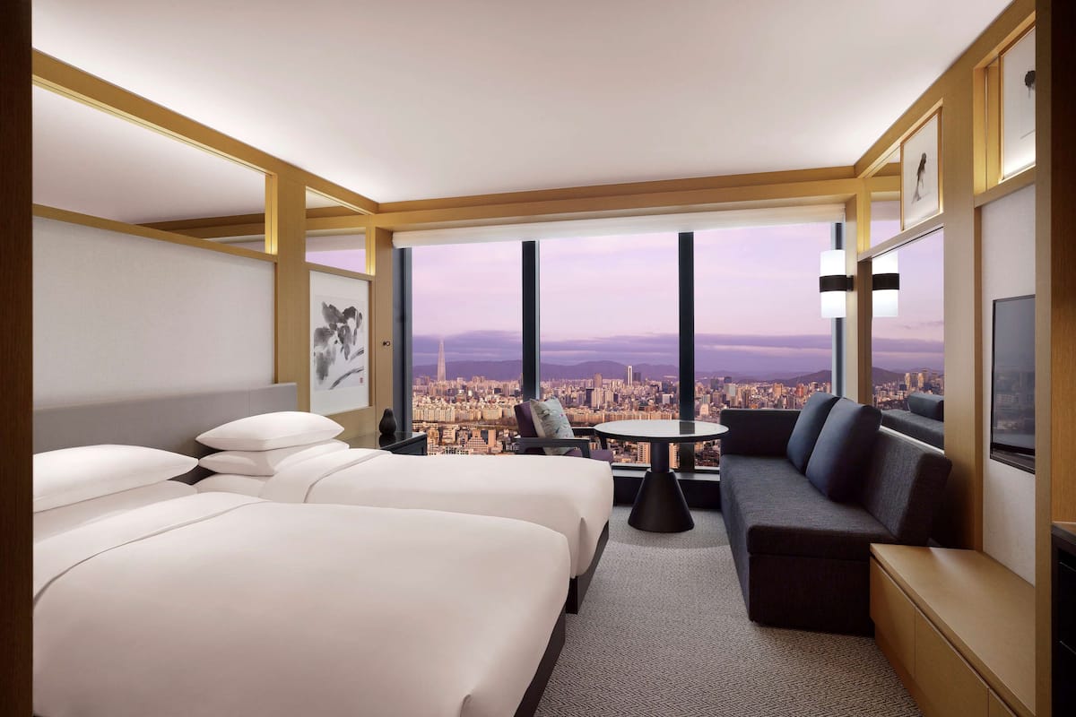 A hotel room with two beds, a sofa, a small table, and a TV. The room has large windows with a city view.