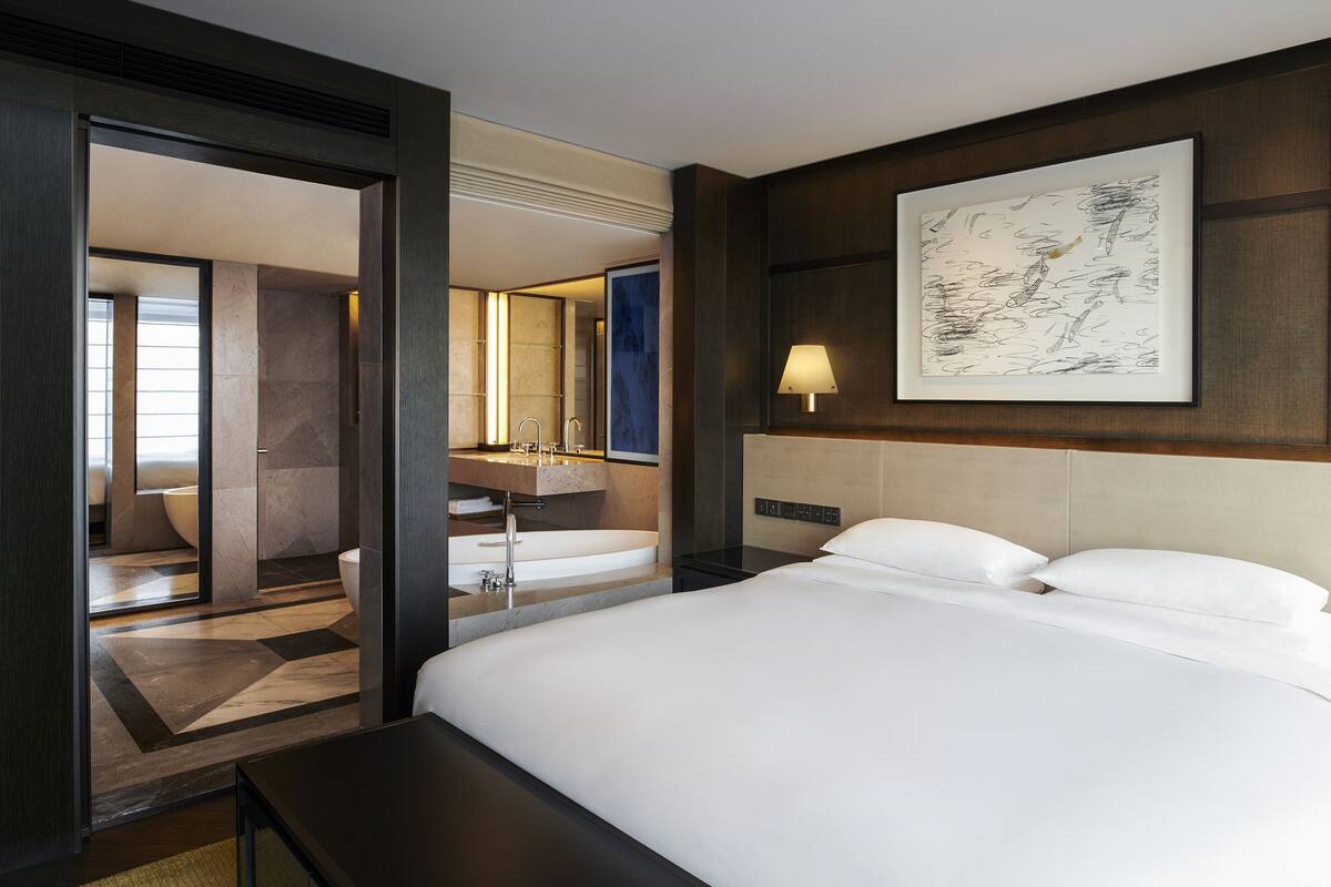 A modern hotel room with a large bed, a bathroom with a bathtub, and a large framed artwork on the wall.