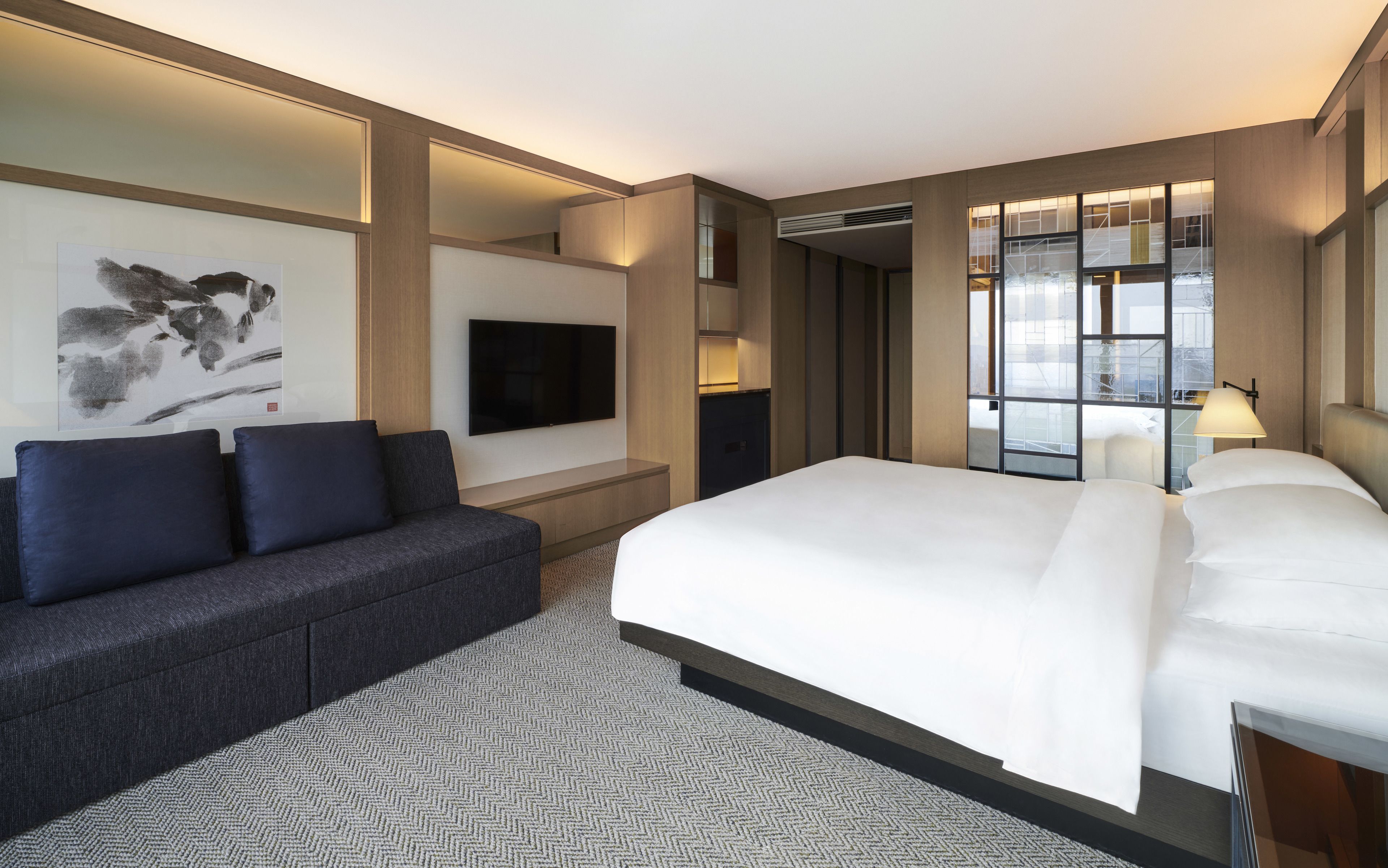 A modern hotel room with a large bed, a sofa, a flat-screen TV, and a city view through large windows.